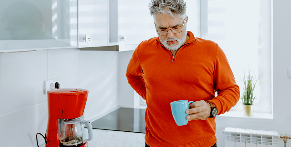 A middle aged man rubs his lower back as if in pain while holding a coffee cup in his other hand.