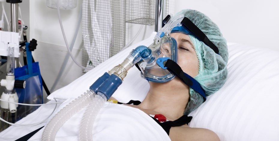 A person is lying in a hospital bed. They are breathing through a respirator, which is strapped to their head. 