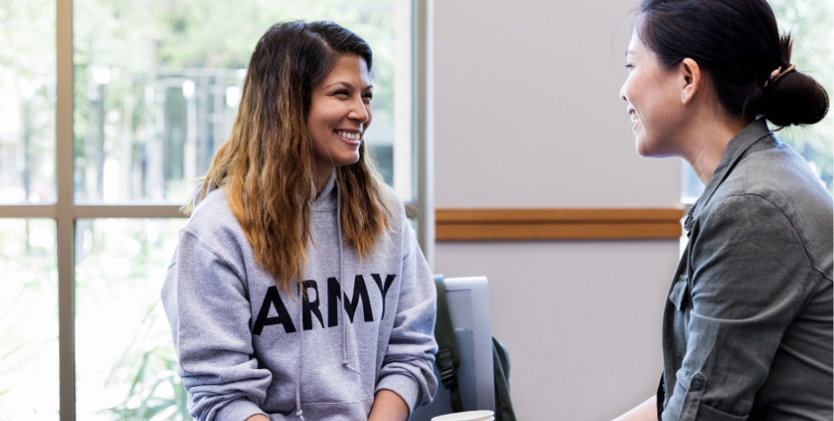 A woman in an army jumper smiles at a group meeting at another woman adopting a caring pose