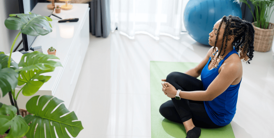 A woman in workout clothes is sitting crosslegged on a yoga mat in a living room. 