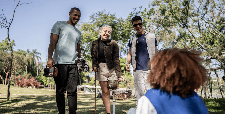 The image shows three people standing up in a park, with a picnic basket and one person sitting down. One of the three people standing has a walking aid and a prosthetic leg. 