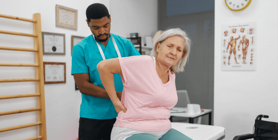 A male physiotherapist examines the back of his female patient, who is sitting on the exam bed. 
