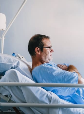 The image is a photo of a middle-aged man in a hospital bed wearing a hospital gown. 