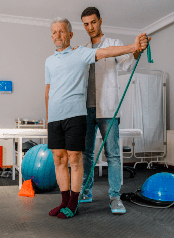 A male physiotherapist stands behind an older man, helping him to do an exercise. 