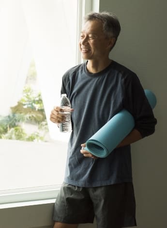 A photo of a man standing by an open window holding a yoga mat and a drink bottle. 