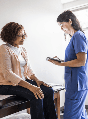 An older woman is sitting on an examination bed. Her physiotherapist is typing something onto a tablet device, 
