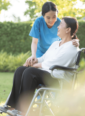 The image shows a female healthcare worker with her hands reassuringly on a woman sitting in a wheelchair
