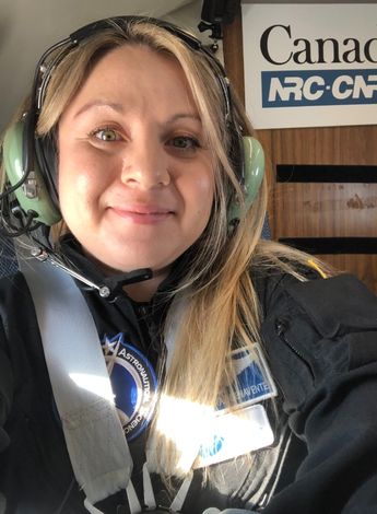 A young blonde woman is strapped into an aircraft seat. She is wearing headphones. 