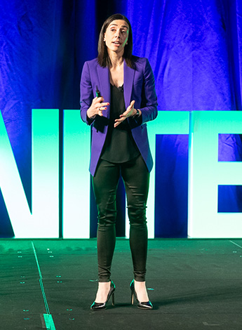 A dark haired women is wearing a purple coat and standing on stage. 