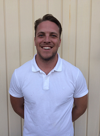 A tanned man wearing a white shirt is standing in front of a cream coloured wall. 