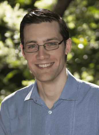 The image is of Dr Sam Manger, a GP who practices lifestyle medicine. He is standing outside in front of green plants and has short dark hair and glasses. 