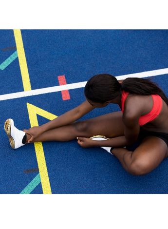 A young black woman in athletics clothing is sitting on an athletics track stretching her legs