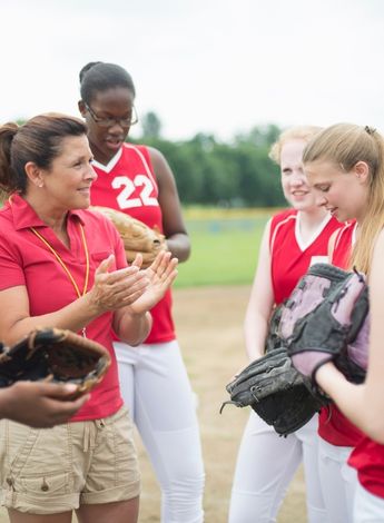 A group of female baseball players stand around their coach as she explains something. 