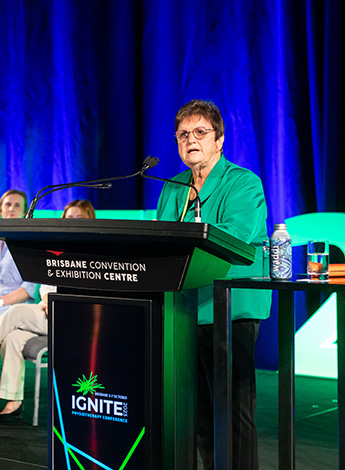 An older woman in a gree suit is standing behind a podium in front of a dark blue curtain. 