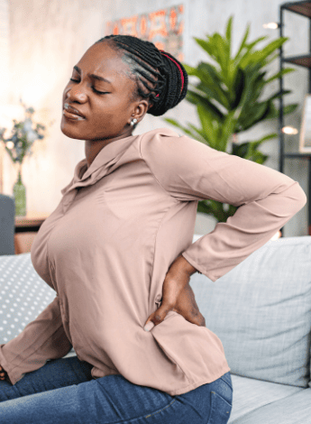 A woman is sitting on a sofa. She is arching her back in pain and her hand is supporting it. 