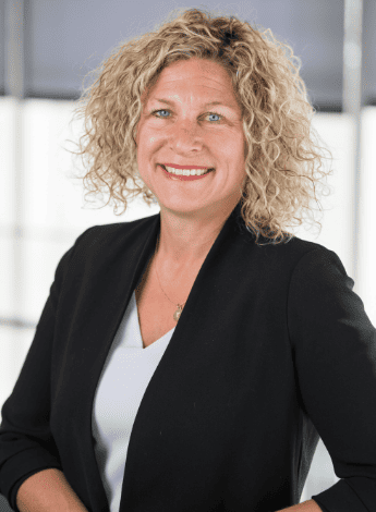The image is physiotherapy researcher Kathryn Schneider. She has blonde curly hair and is wearing a black blazer over a white shirt. 