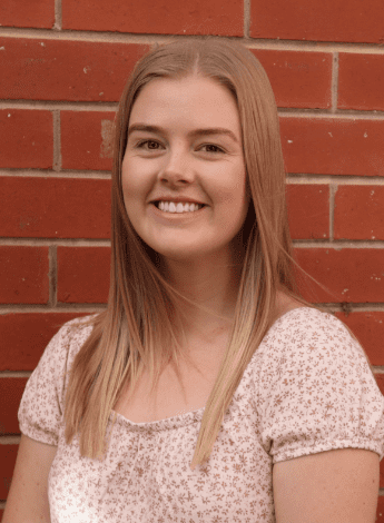 A woman with long blonde hair is standing in front of a red brick wall. 