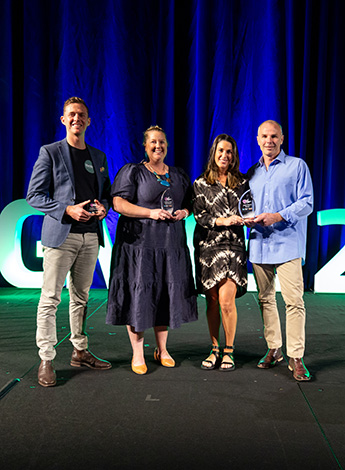 Two men and two women stand on a stage in front of a blue curtain. They are all holding awards. 