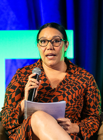 A woman wearing an orange shirt speaks to an audience. 