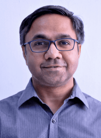 The image is of an older Indian man with dark hair and glasses