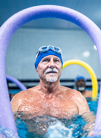 An older man is in a pool holding up a pool noodle. 