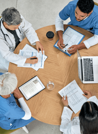 The image shows a group of health professionals sitting around a table in a meeting. The photo is taken from above, looking down on the meeting.