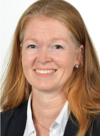 The image is of a woman with long strawberry blonde hair. She is wearing a white shirt and a dark blazer. 