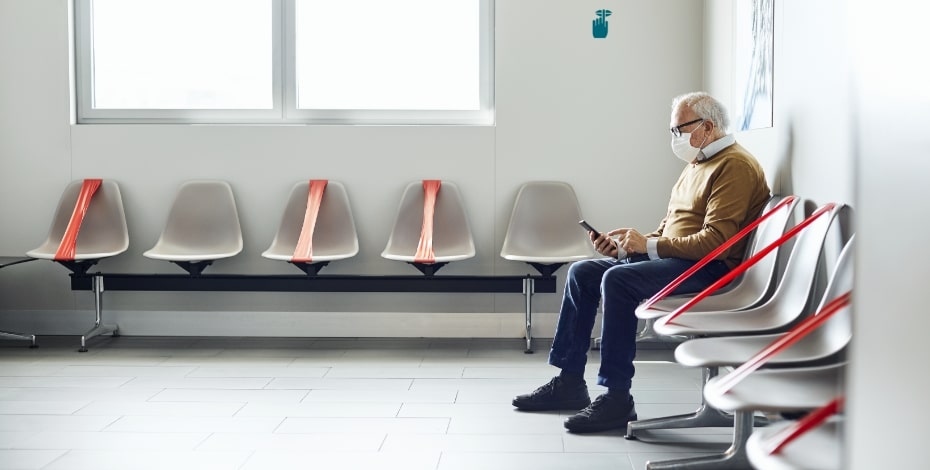 Elderly man sits in a waiting room with chairs taped off. 