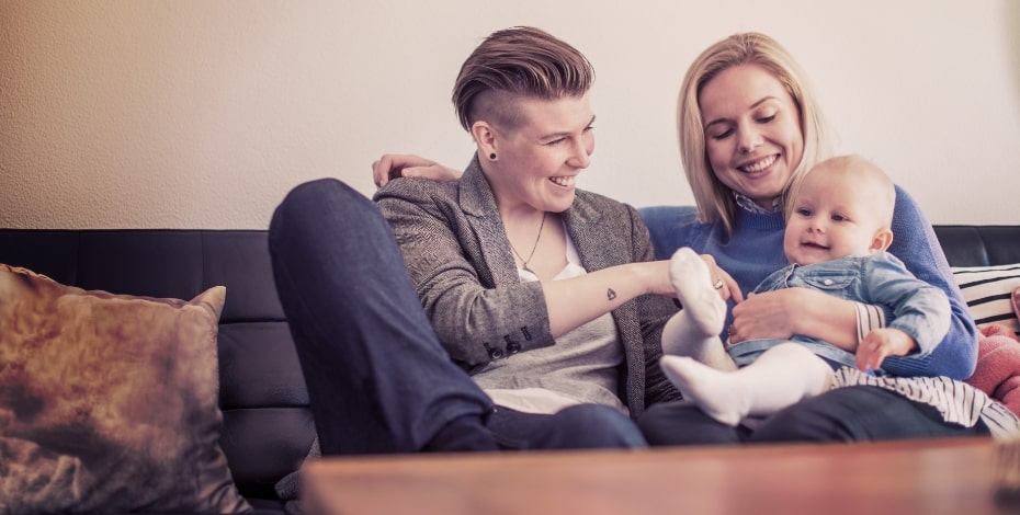 A family sitting on the couch: two mums cuddling their baby.