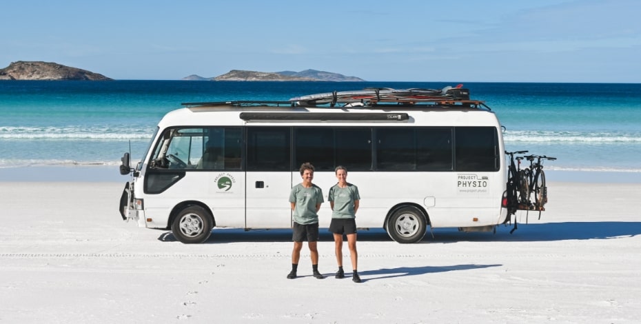 Two physios stand in front of a large van on the beach.