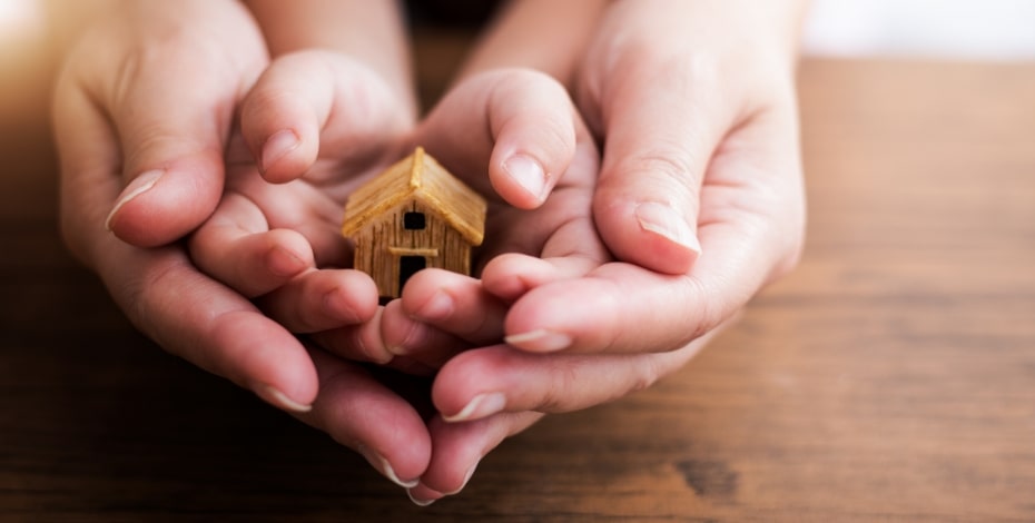 Adult hands around child hands holding a tiny wooden house.