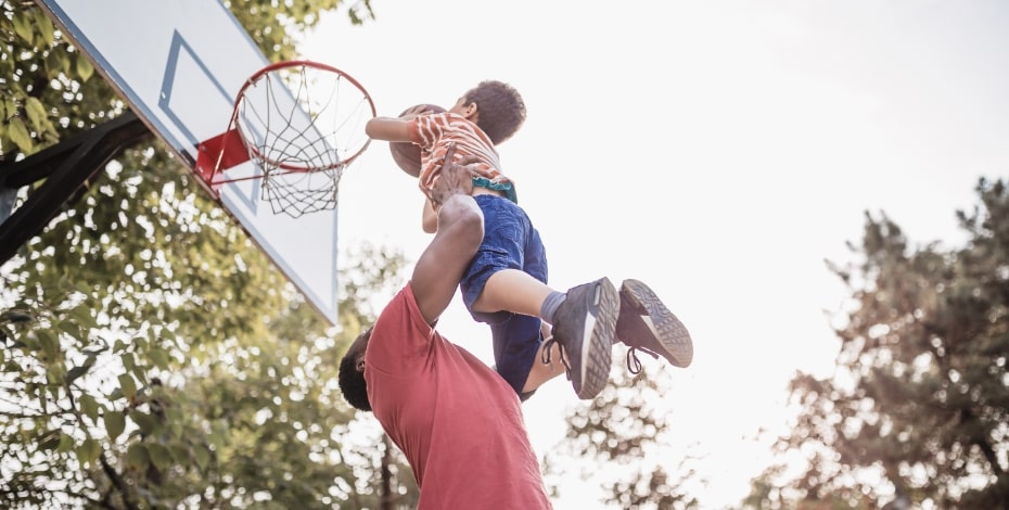 A man lifting a male child in the air and up to a basketball hoop on a pole.