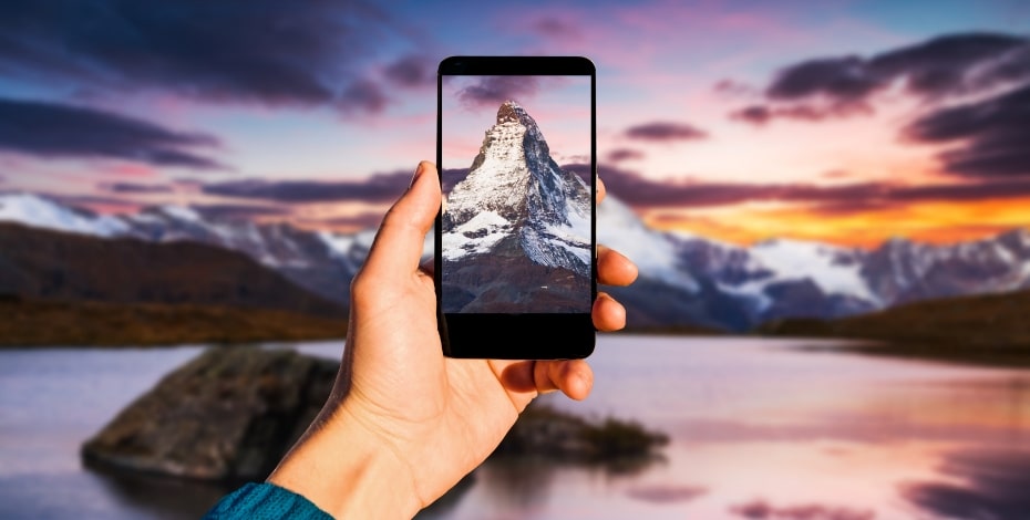 A hand holding a mobile phone up to photograph a sunset.