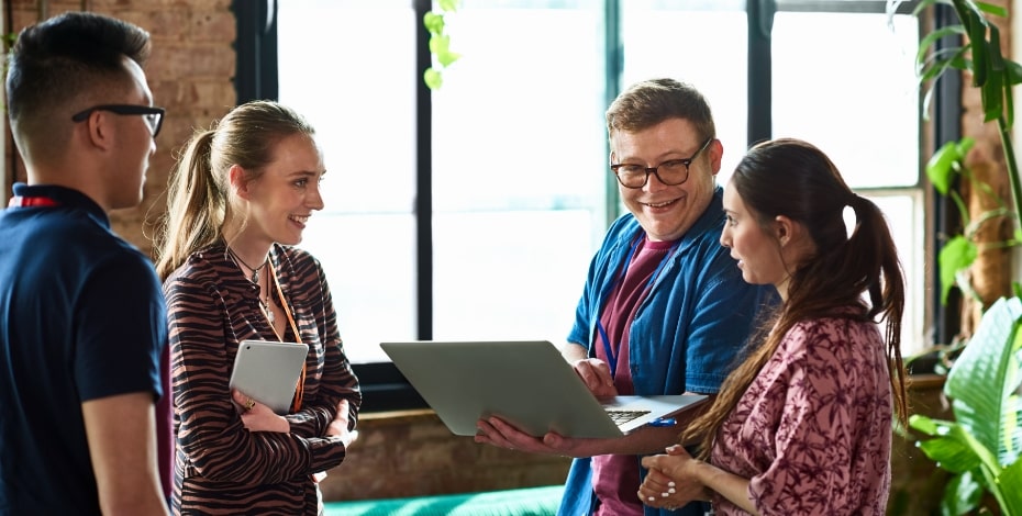 A group of smiling employees have a discussion looking at a laptop. 