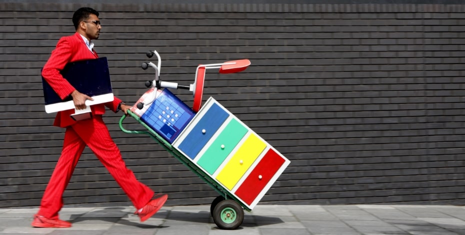 A man in a red suit pushes a two-wheel trolley with office furniture and equipment stacked on it.