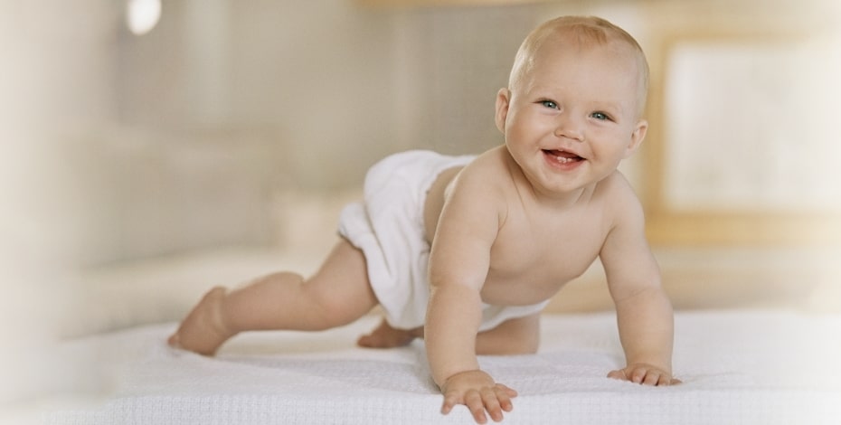 A smiling baby wearing a nappy crawls on a white blanket. 