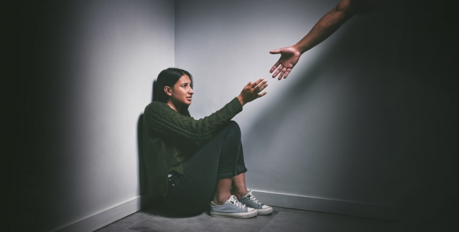 A woman huddles in a corner looking sad with someone reaching out a hand to her.
