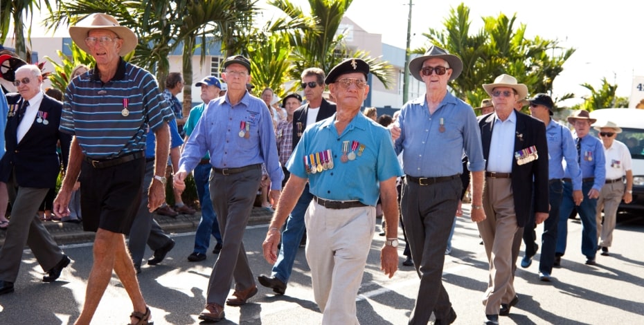 A group of ageing military personnel in neat casual attire walks together in a parade.