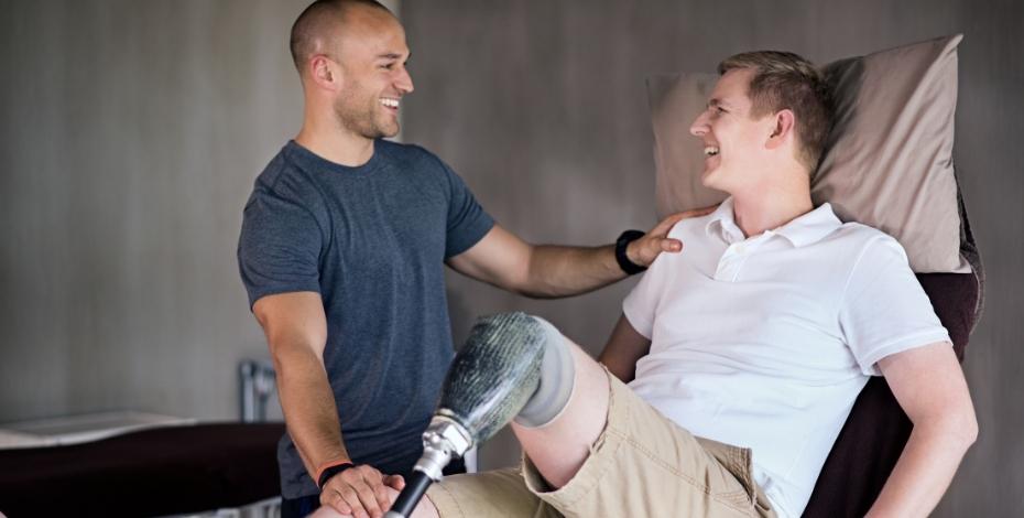 A male physiotherapist is standing up, smiling and touching the shoulder of a male patient who is sitting upright on a treatment table, looking at, and smiling back at the physiotherapist. The patient has a prosthetic leg.