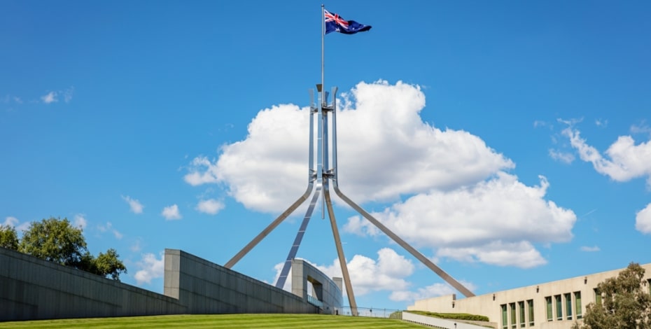 A view of Parliament House from the base of the hill, with 'stripy' newly mowed grass, a building on one side, a concrete stepped wall on the other and at the top an enormous multi-pronged flagpole.