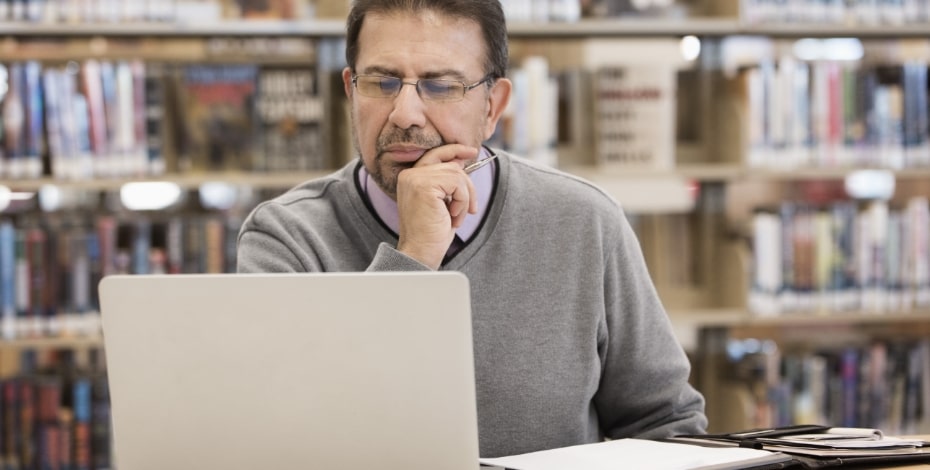 A man is sitting in front of library bookshelves looking at the screen of his laptop computer.