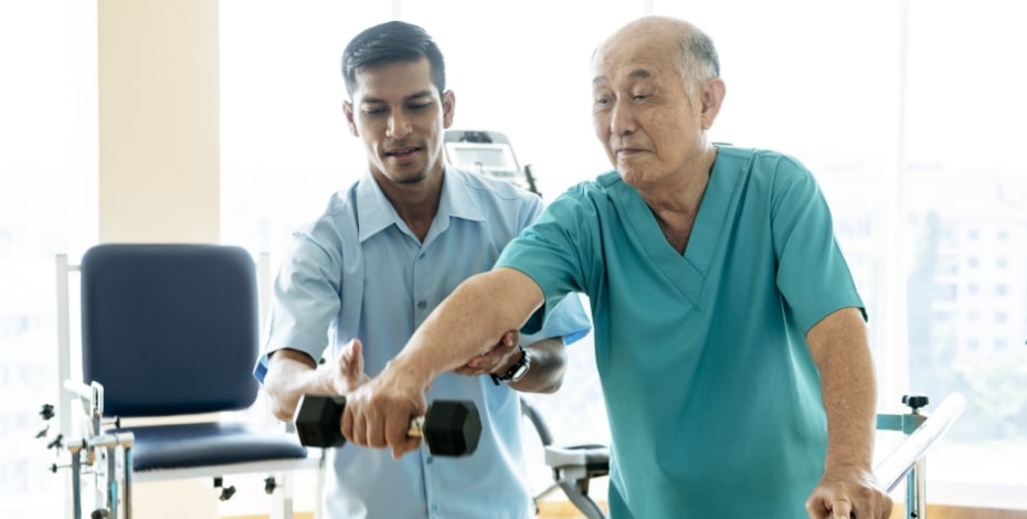 A male physiotherapist in a blue shirt helps an elderly Asian man to lift a hand weight