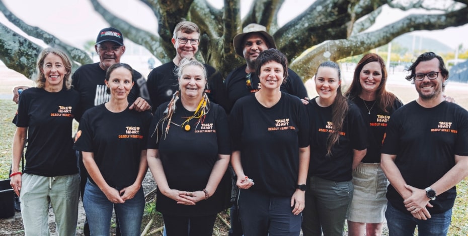 A group of ten men and women wearing black T-shirts with the Deadly Heart Trek logo over the heart stand in front of a large tree. 