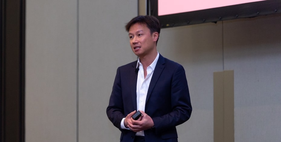 An Asian man in a suit stands in front of a screen at a conference