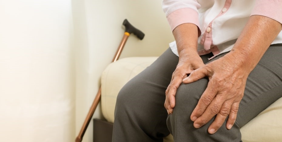 A person in a white shirt and grey trousers sits on a bed holding their left knee. A walking stick rests against the bed. 