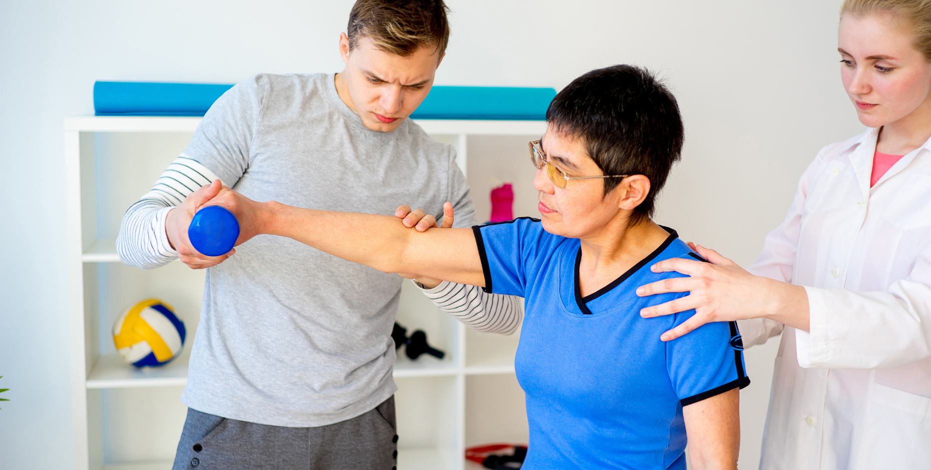 An older patient is lifting a small weight in the right hand with two clinicians correcting and monitoring their technique