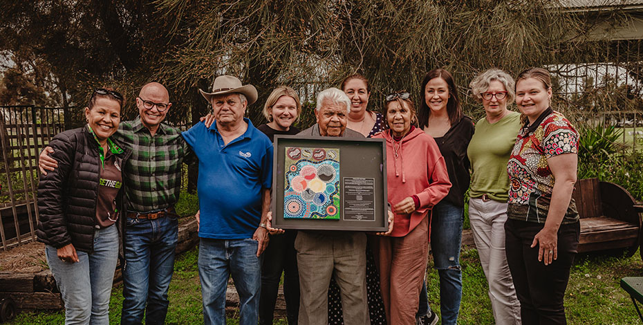 A group of mean and women are standing around an Aboriginal man holding an Aboriginal artwork. Some of them are also Aboriginal people.  