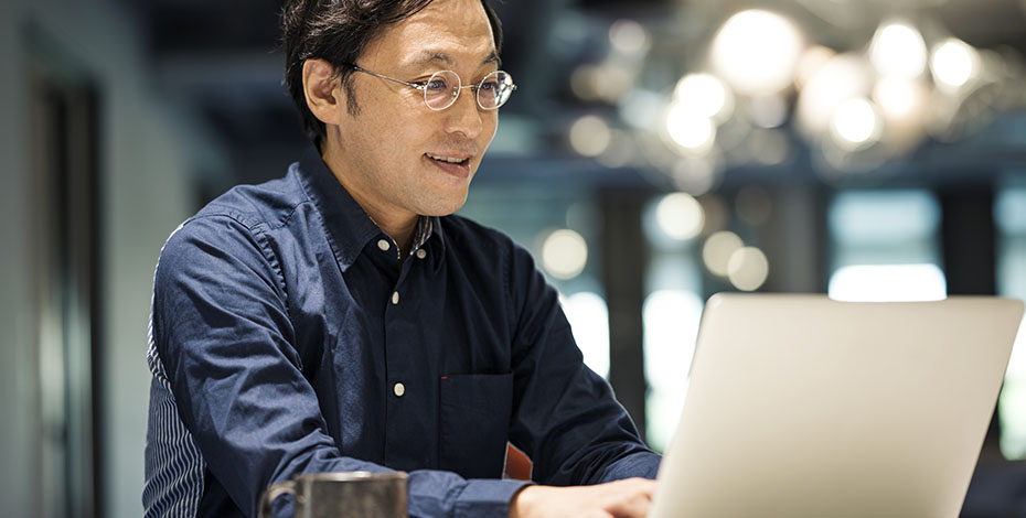 A happy looking man in a blue shirt works at a laptop with a coffee next to him.