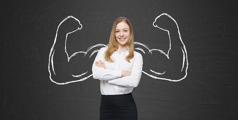 A woman stands in front of a chalkboard with muscly arms flexing drawn on the board behind her.