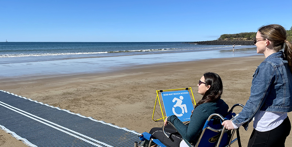 A woman is pushing another woman in wheelchair down a grey sand mat over the sand towards the water at the beach. The wheelchair has special  fat tires for sand.